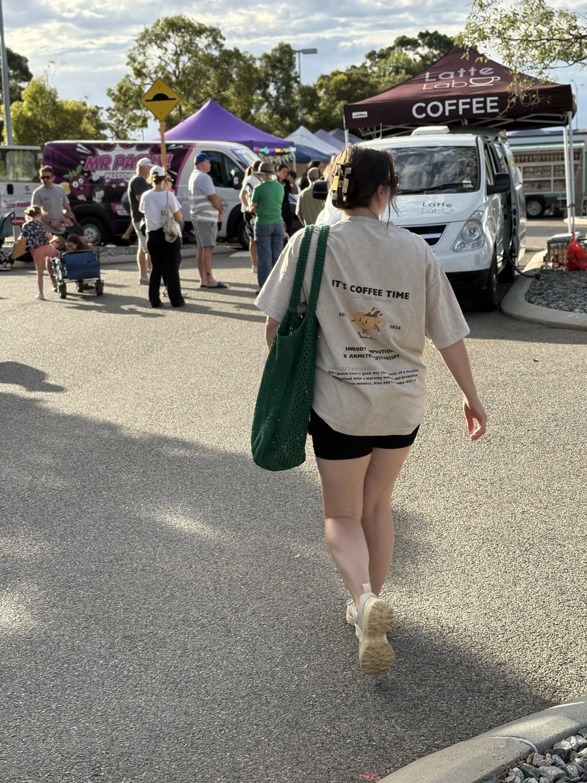 A young woman with dark hair tied with cloth, wearing a beige t-shirt with printed text and black shorts, walking past a busy outdoor market setup with tents and vendors, and a white van in the background, on a sunny day.