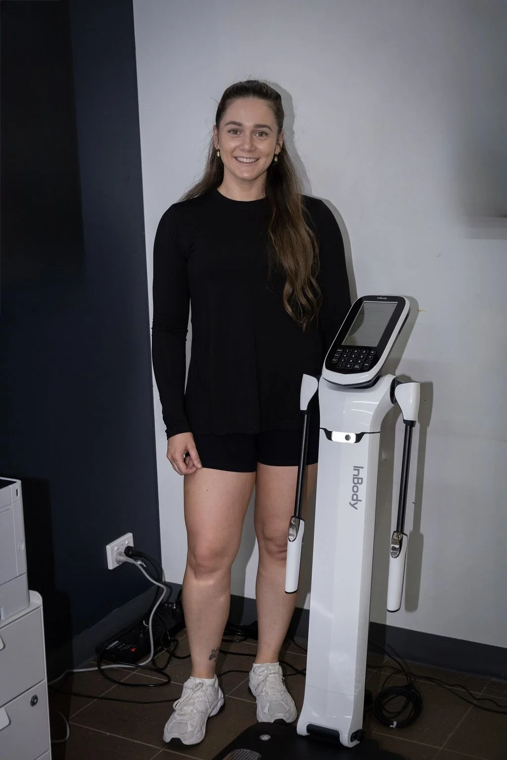 A woman in black workout clothing standing next to a body composition analyzer in a gym or fitness center.