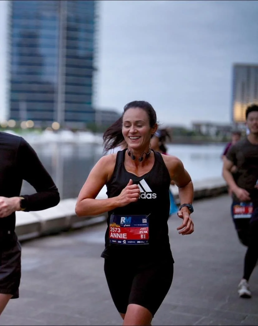 A woman running in a marathon by a river with tall buildings in the background, wearing a black athletic outfit and a race bib that reads 'Annie'.