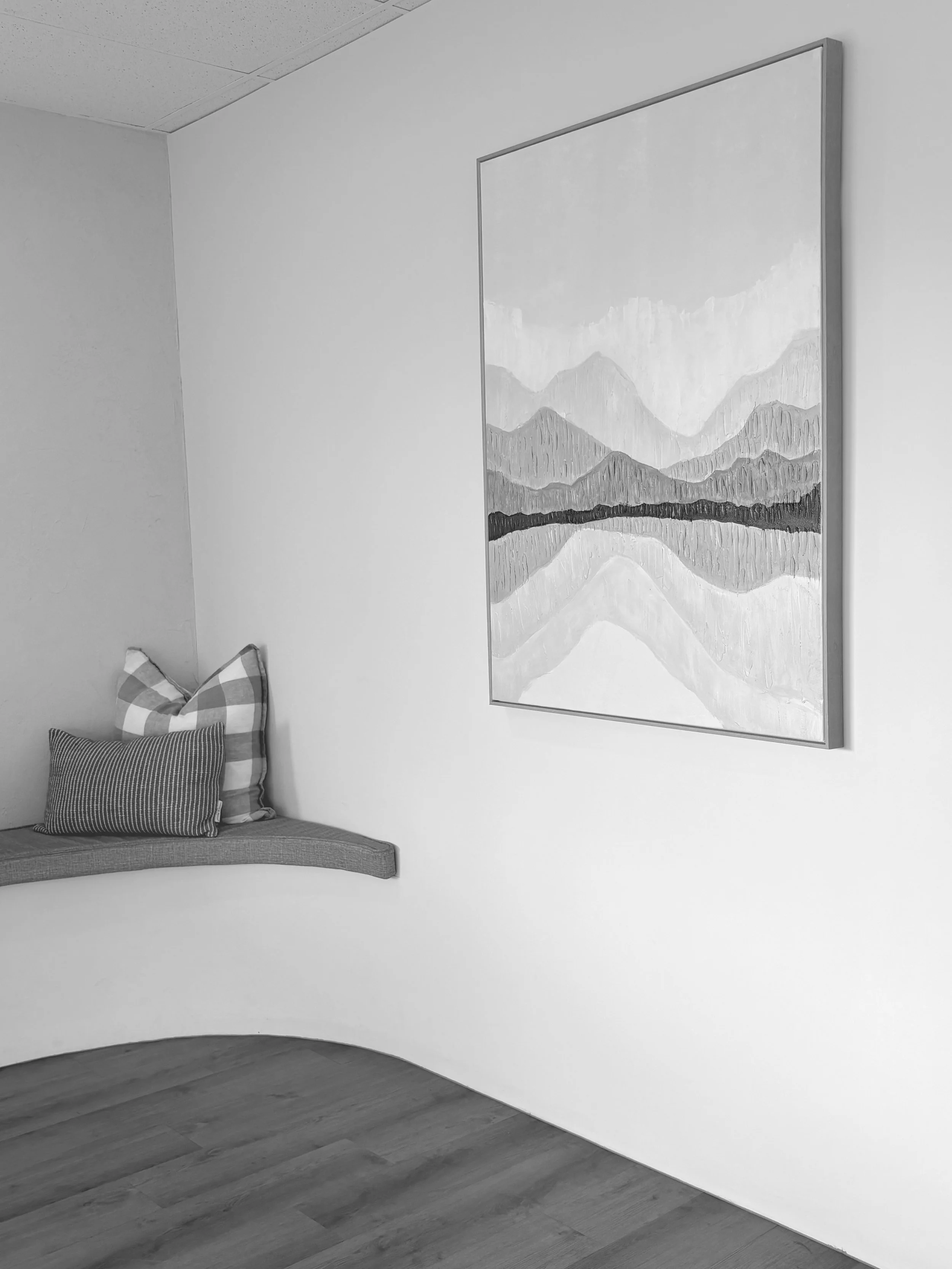 Black and white photograph of an interior space with a white wall, wooden flooring, a framed painting of mountains and a lake, and a corner bench with pillows.