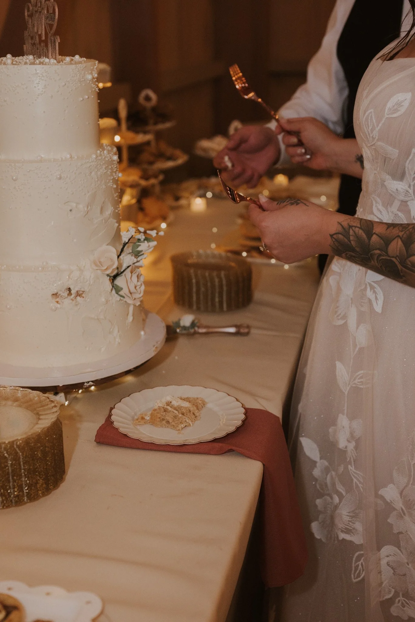 People cutting a wedding cake decorated with white flowers and pearls at a wedding reception, with a table of desserts in the background.