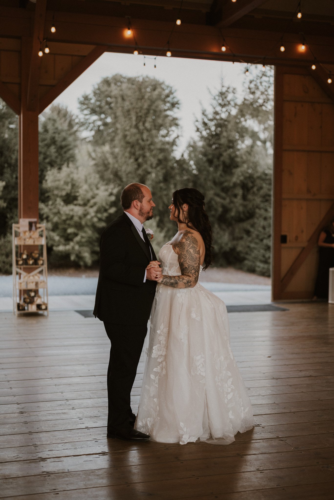 A bride and groom holding hands and looking at each other during their wedding ceremony, inside a wooden venue with outdoor scenery in the background.