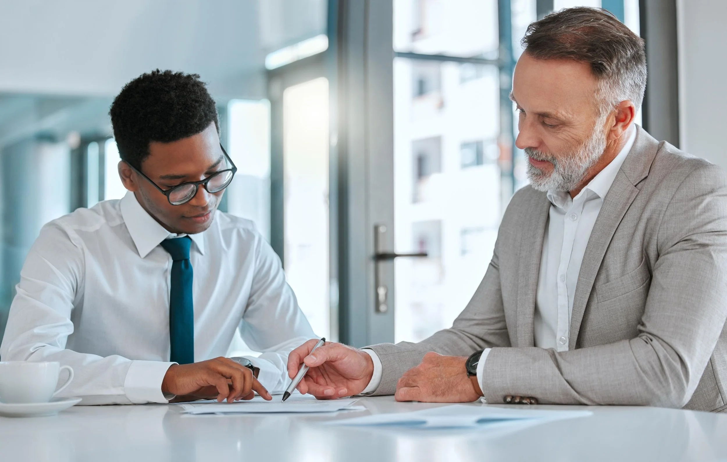 Two men in business attire sitting at a table in a modern office, looking at documents and discussing.