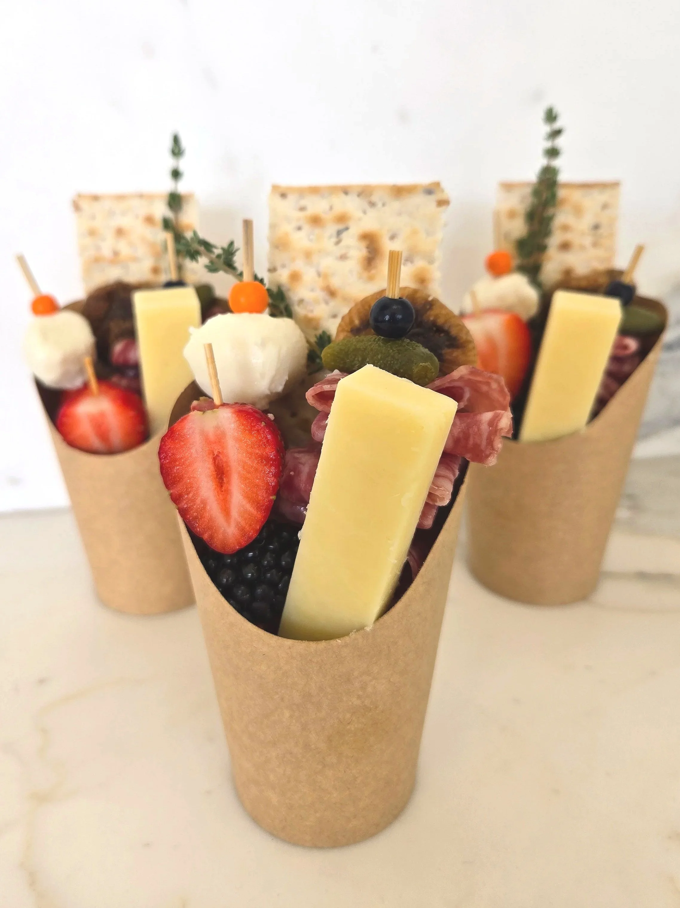 Two paper cones filled with various snacks and garnished with strawberries, blackberries, and herbs, arranged on a white surface.
