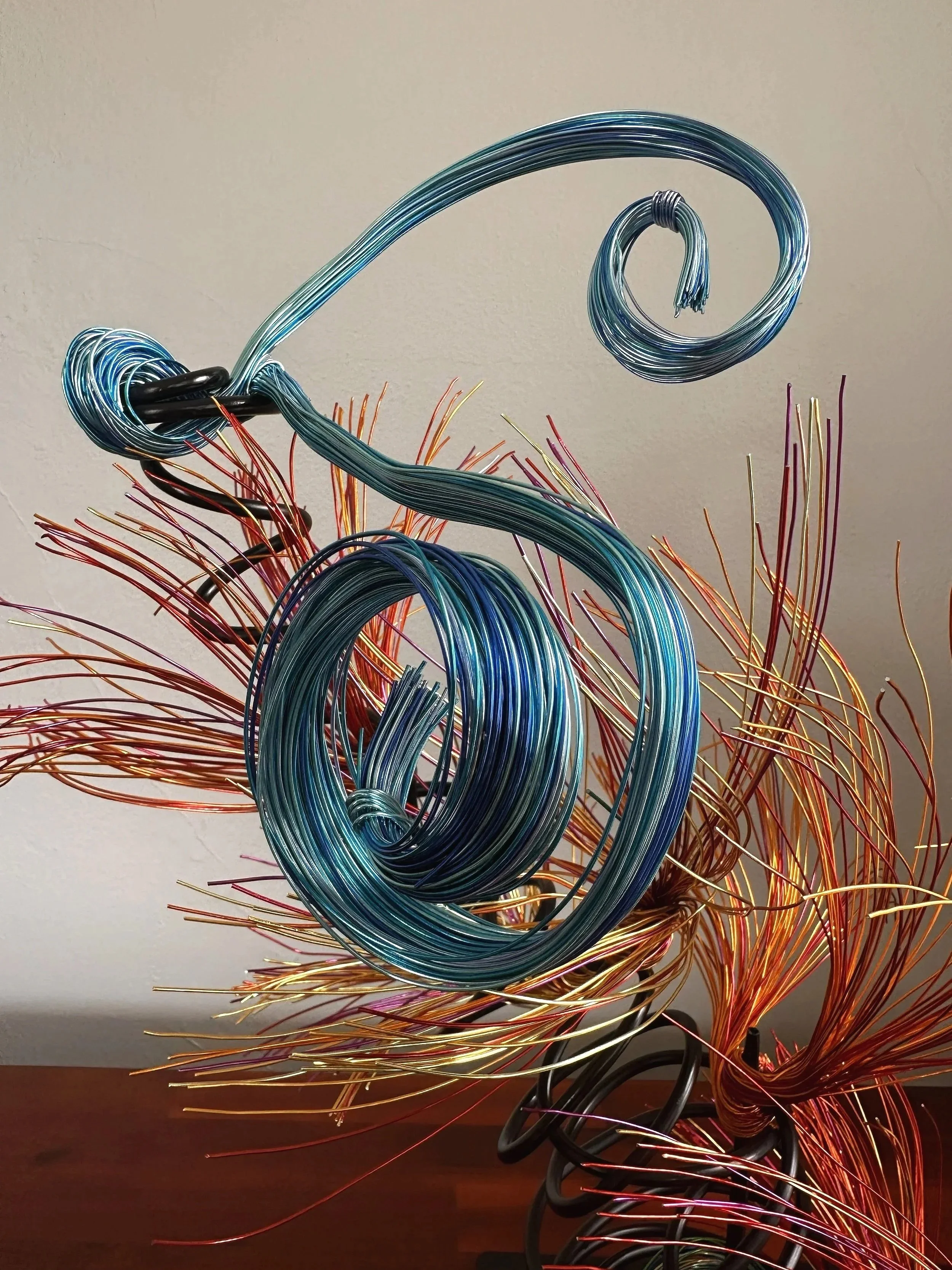 Title: "Flowing" View from front 
Dimensions: 1' × 1' × 2'
 Red and Orange wire rises from a grounded base into a quiet, upward motion — a gesture shaped slowly by hand, carrying both volcanic tension and blue wave lift
