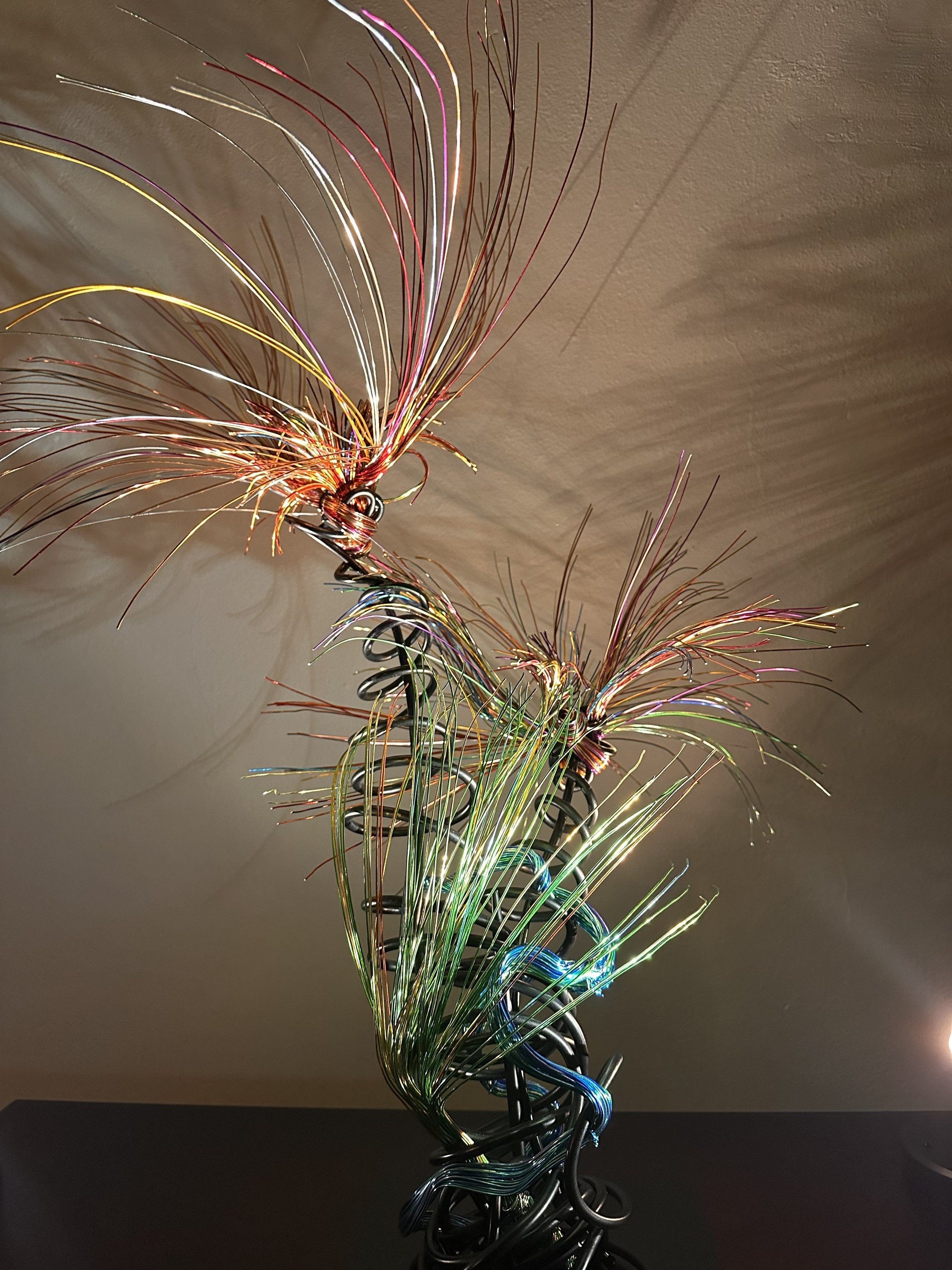 Title: Luminous. View from front. Spot Light
Dimensions:  2' × 2 '× 2' 8"
Fine wires flare outward like petals deciding to open, catching the light in shifting tones of copper, ember, and moss. The piece holds the stillness of a plant just on the edg