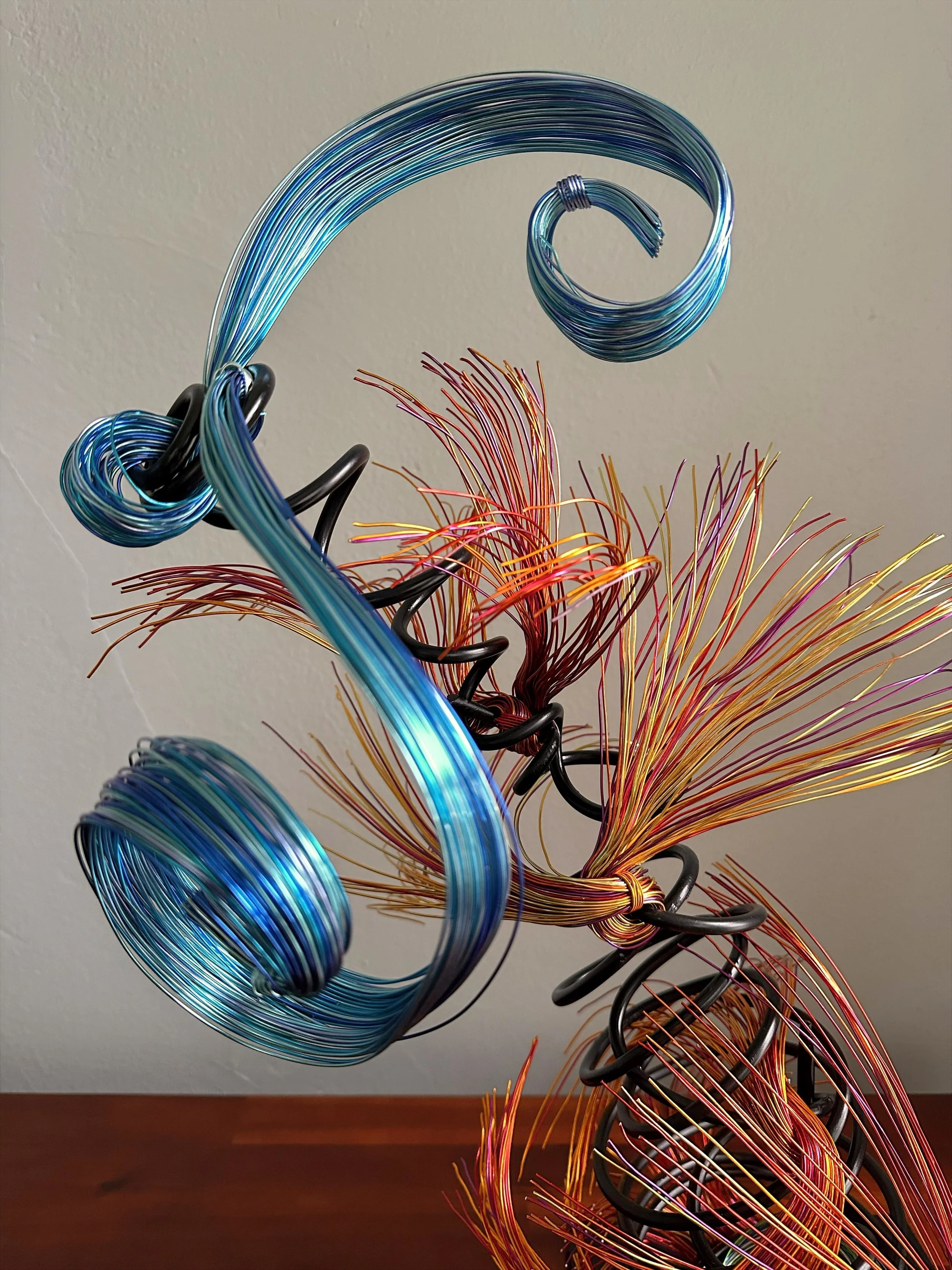 Title: "Flowing" View from front 
Dimensions: 1' × 1' × 2'
 Red and Orange wire rises from a grounded base into a quiet, upward motion — a gesture shaped slowly by hand, carrying both volcanic tension and blue wave lift