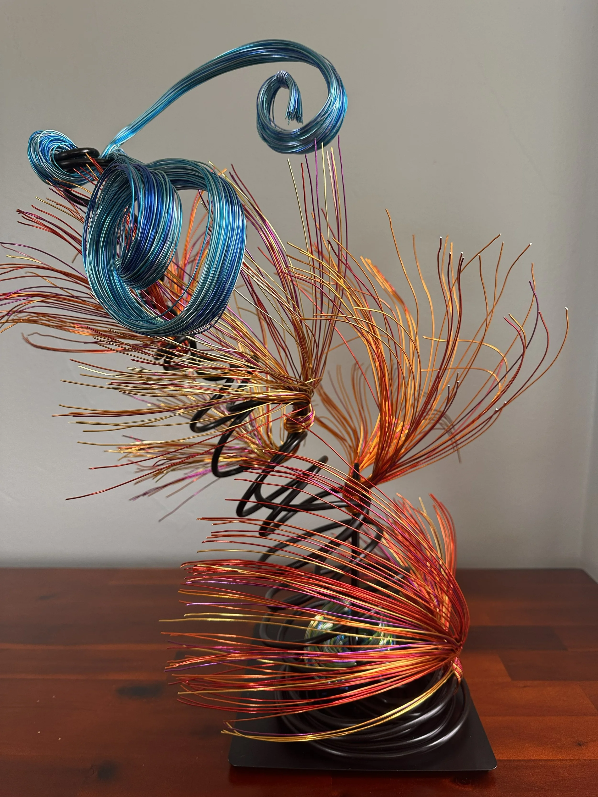 Title: "Flowing" View from front. Detail 
Dimensions: 1' × 1' × 2'
 Red and Orange wire rises from a grounded base into a quiet, upward motion — a gesture shaped slowly by hand, carrying both volcanic tension and blue wave lift