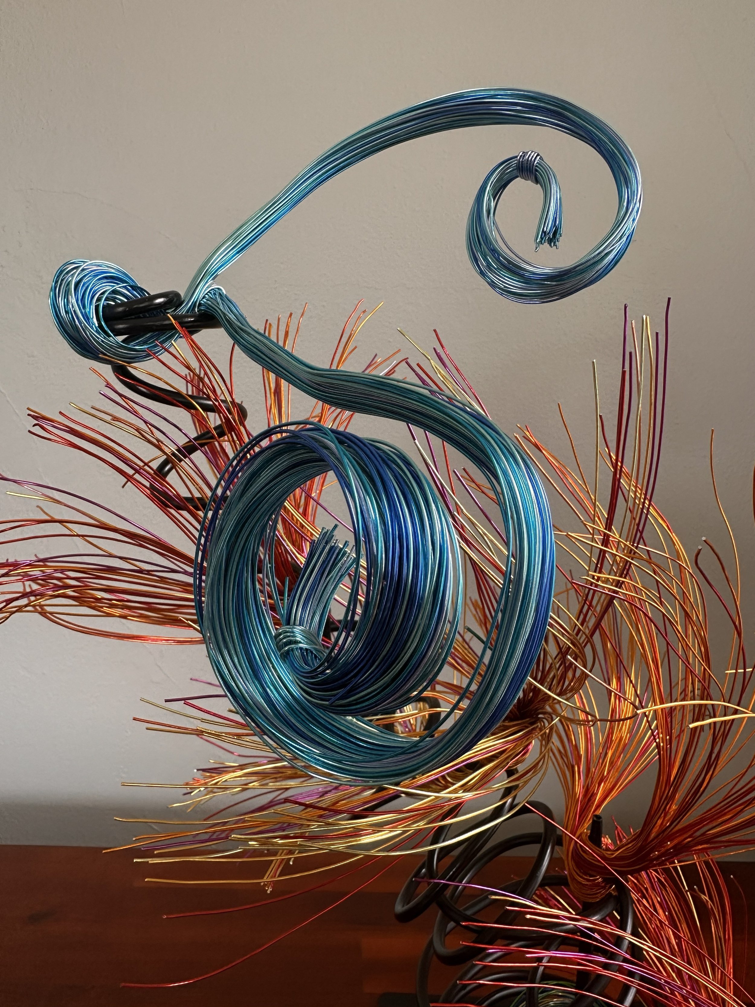 Title: "Flowing" View from front 
Dimensions: 1' × 1' × 2'
 Red and Orange wire rises from a grounded base into a quiet, upward motion — a gesture shaped slowly by hand, carrying both volcanic tension and blue wave lift