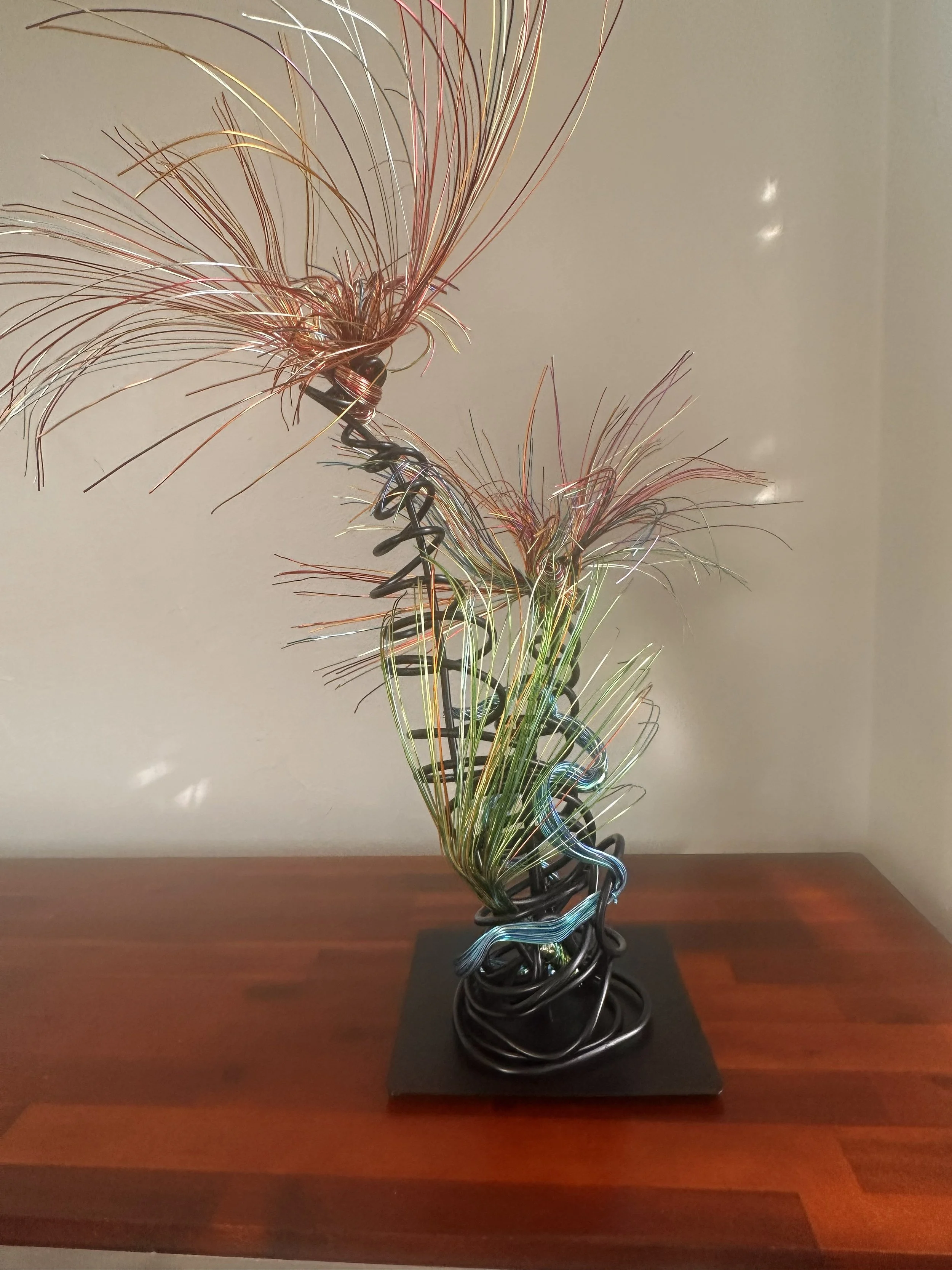 Title: Luminous.  View from front
Dimensions:  2' × 2 '× 2' 8"
Fine wires flare outward like petals deciding to open, catching the light in shifting tones of copper, ember, and moss. The piece holds the stillness of a plant just on the edge of becomi