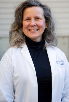 Smiling woman with wavy gray hair wearing a white lab coat and a black turtleneck, standing outdoors in front of a beige wall. Beauty Period Aesthetics in Wasco, IL offers Botox, dermal fillers, microneedling, wellness injections, and weight loss