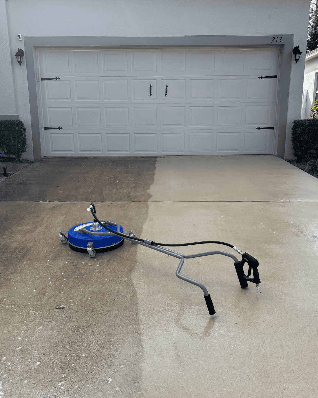 A concrete driveway being power washed, with half cleaned and half dirty section, in front of a white garage door with house number 213.