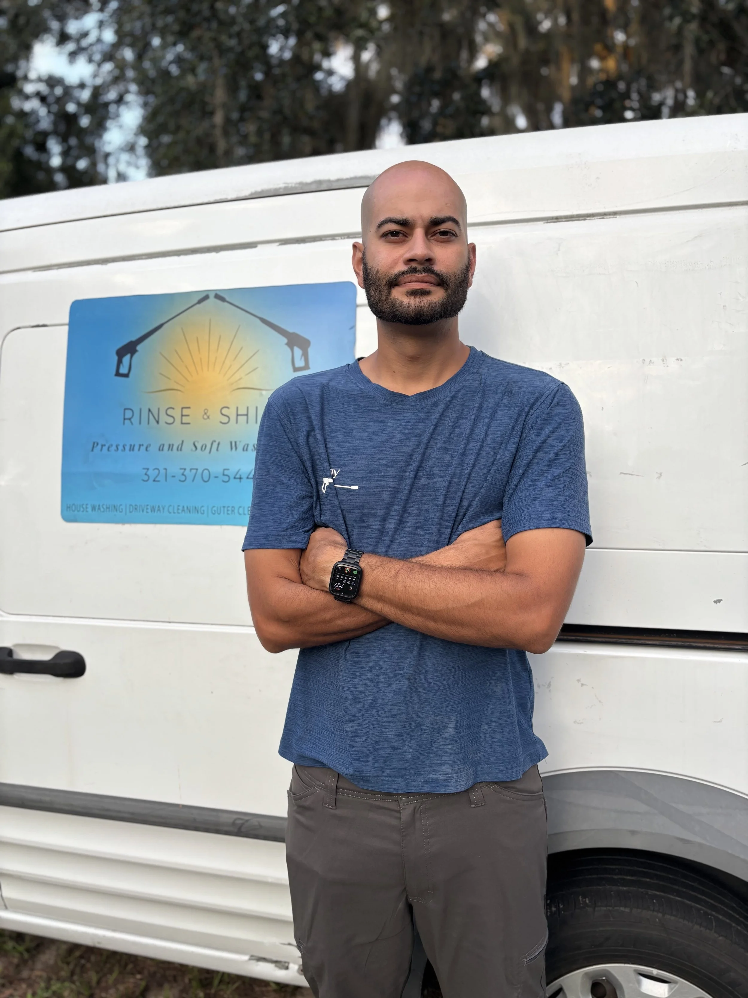 A man with a bald head and beard standing with crossed arms in front of a white van with a sign that reads 'Rinse & Shine Pressure and Soft Wash.' He is wearing a blue T-shirt and gray shorts, and is outdoors with trees in the background.