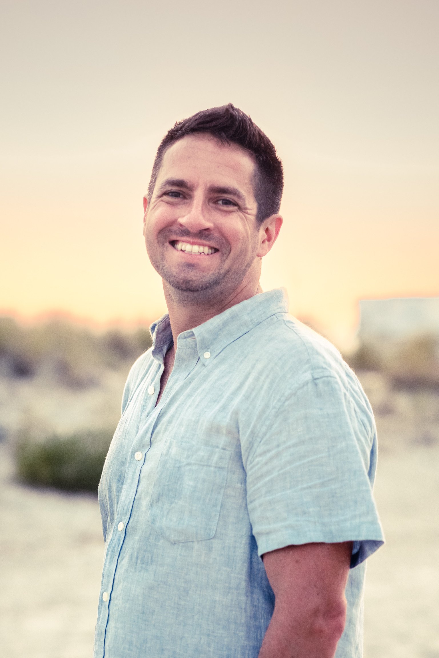 Smiling man in a light blue shirt outdoors at sunset with a blurred beach background.