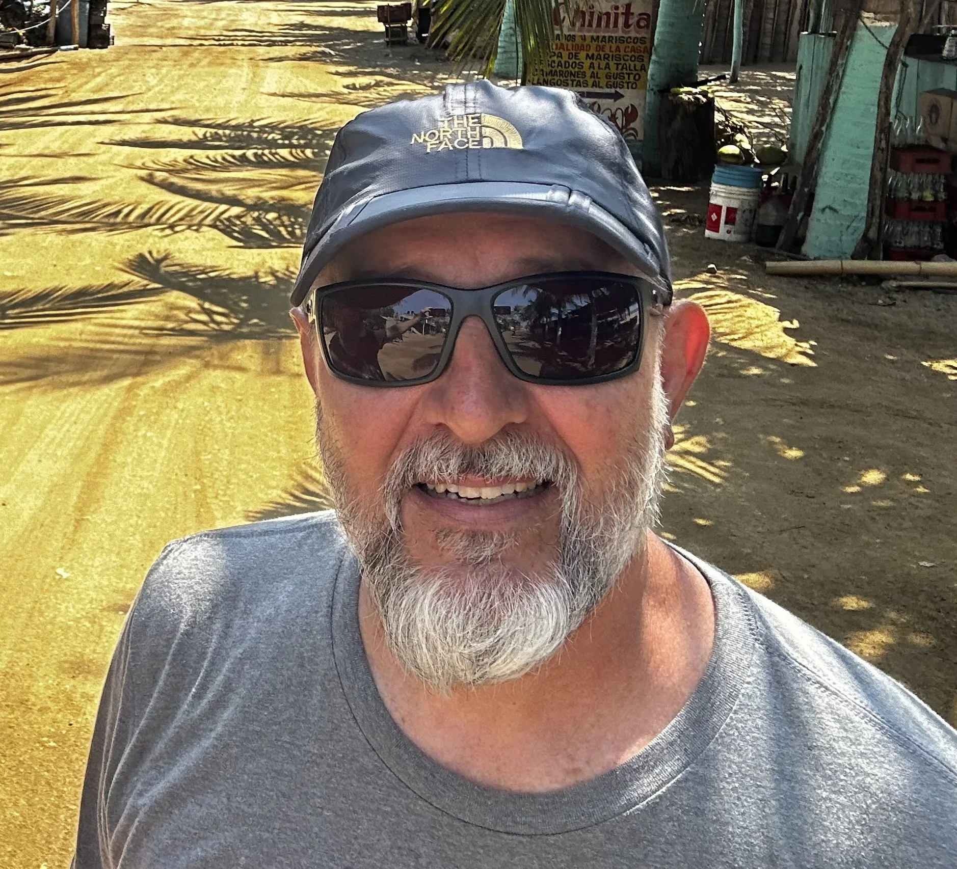 A smiling man with a gray beard wearing sunglasses, a gray t-shirt, and a black cap outdoors on a sunny day, with shadows of palm trees on the ground behind him.