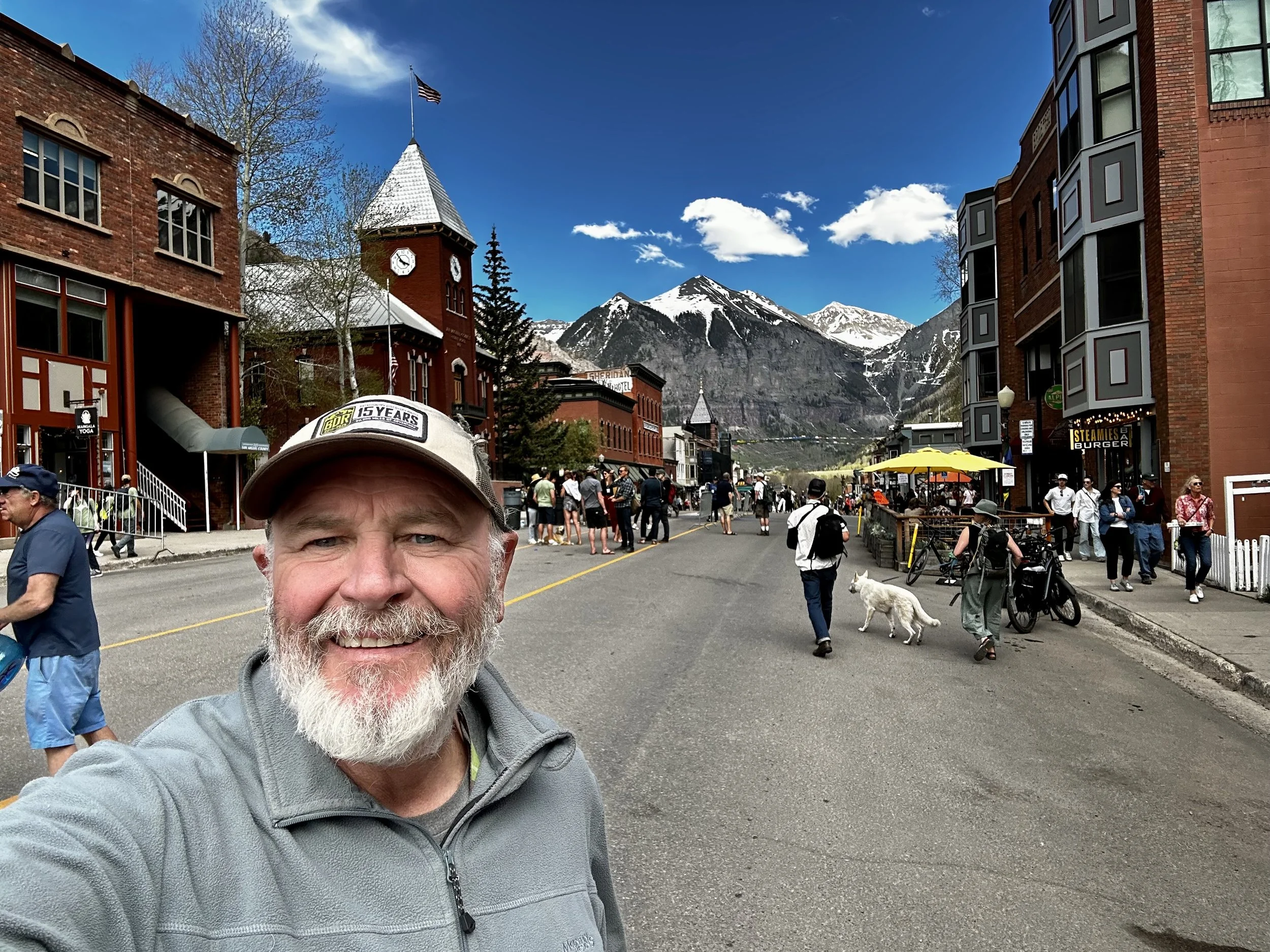 Smiling man taking a selfie on a bustling mountain town street with crowd, colorful buildings, and snow-capped mountains in the background.