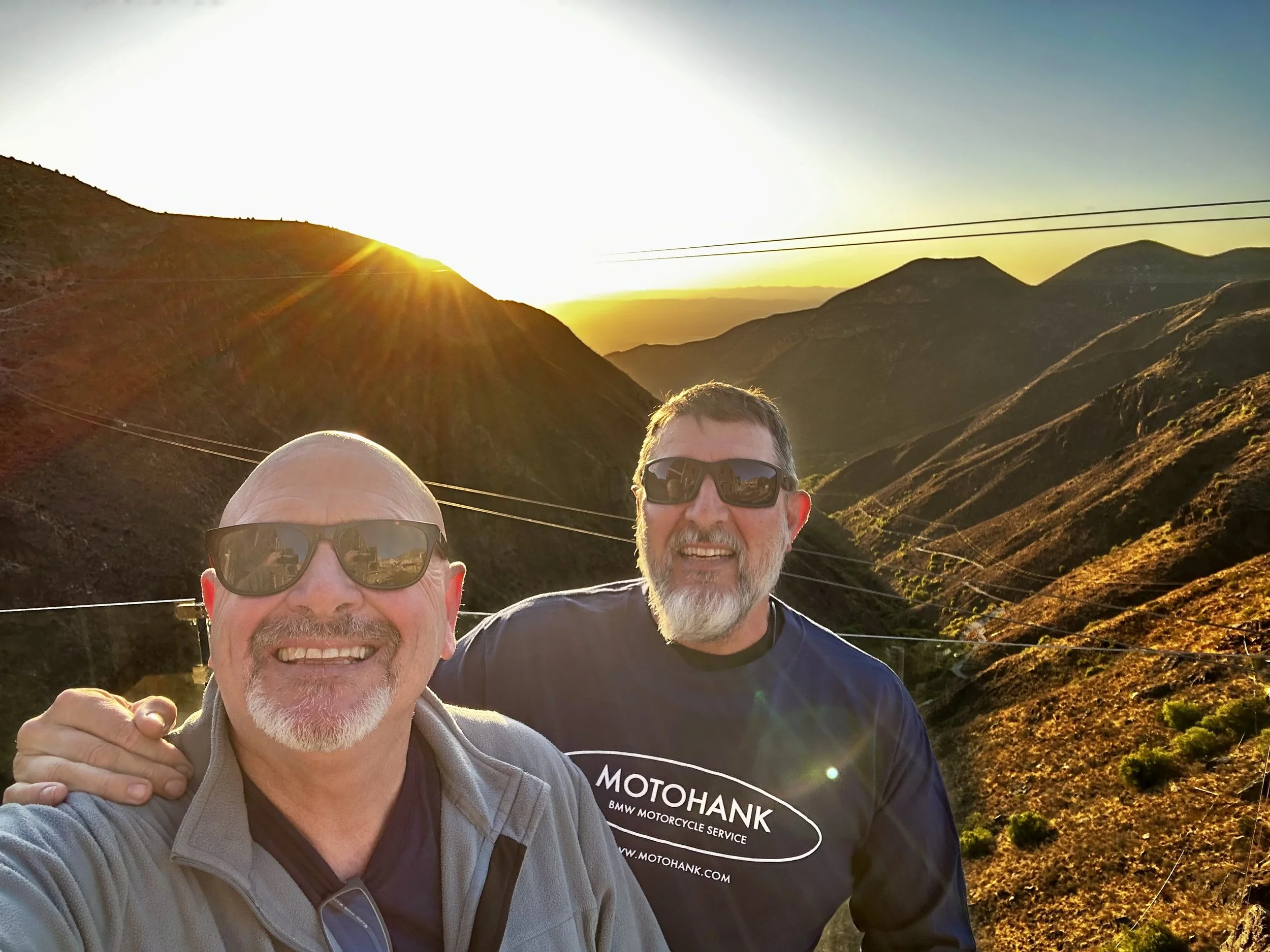 Two men wearing sunglasses smiling for a selfie in a mountainous landscape during sunset, with hills and power lines in the background.