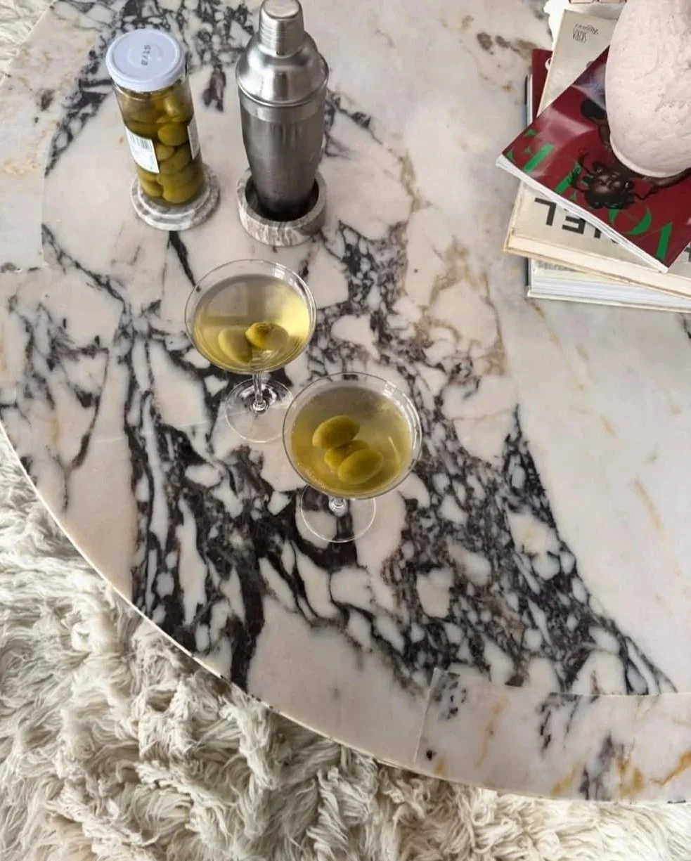 Two glasses of green olives in liquid on a marble table, with a jar of green olives, a cocktail shaker, and magazines nearby.