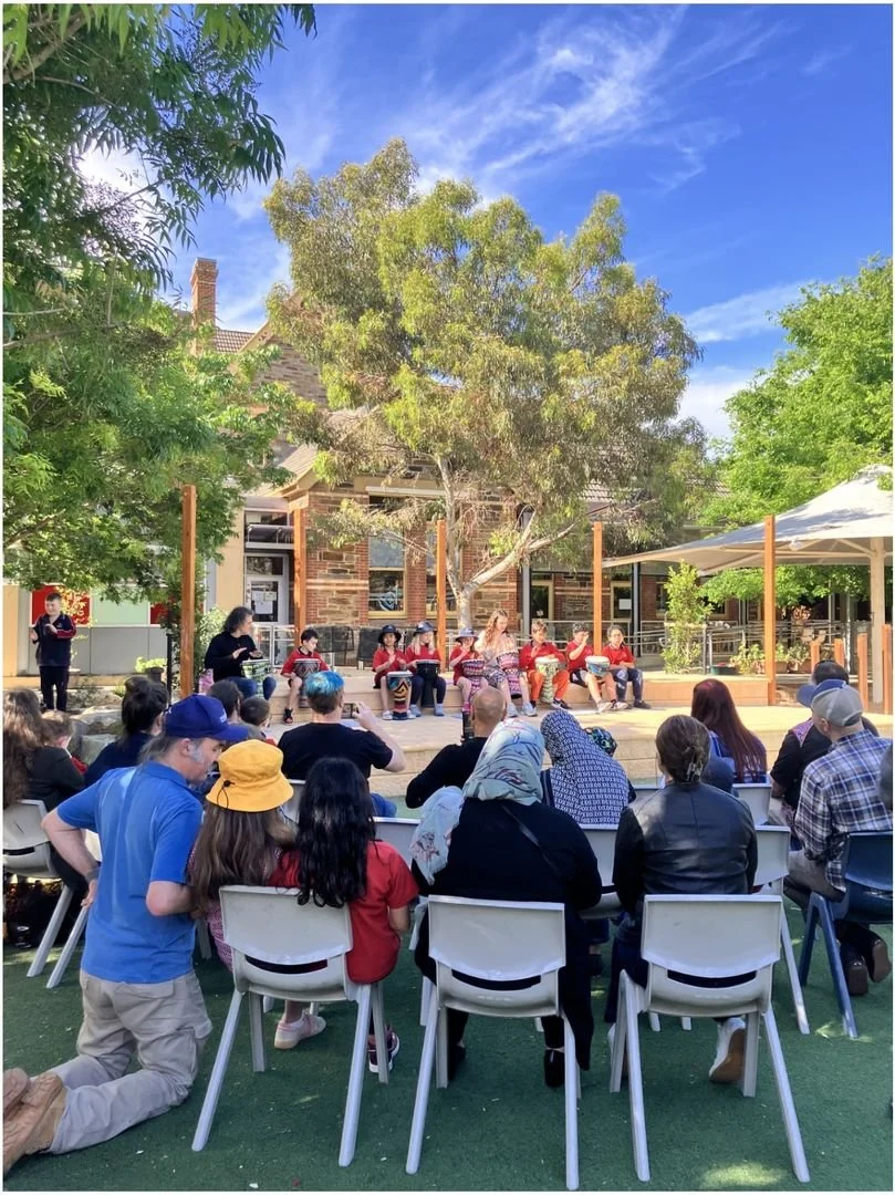 Outdoor gathering of people watching a performance or presentation on a small stage under a large tree, with a brick building in the background and a clear blue sky.