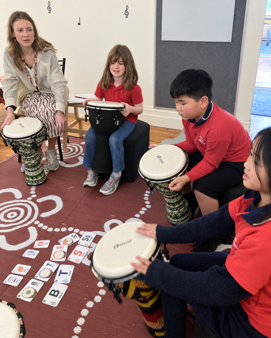 A music class with a teacher and children playing drums and using word cards on the floor.