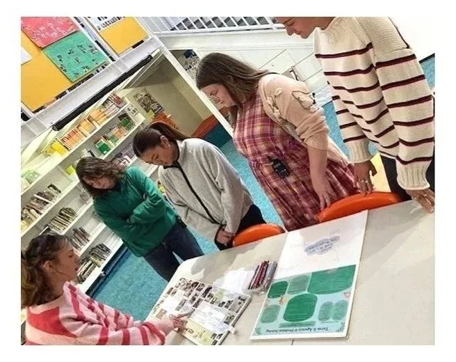 A group of five friends or classmates gathered around a table in a library or classroom, looking at a magazine or book with colorful pages, while one girl is seated and appears to be explaining or showing something to the others.