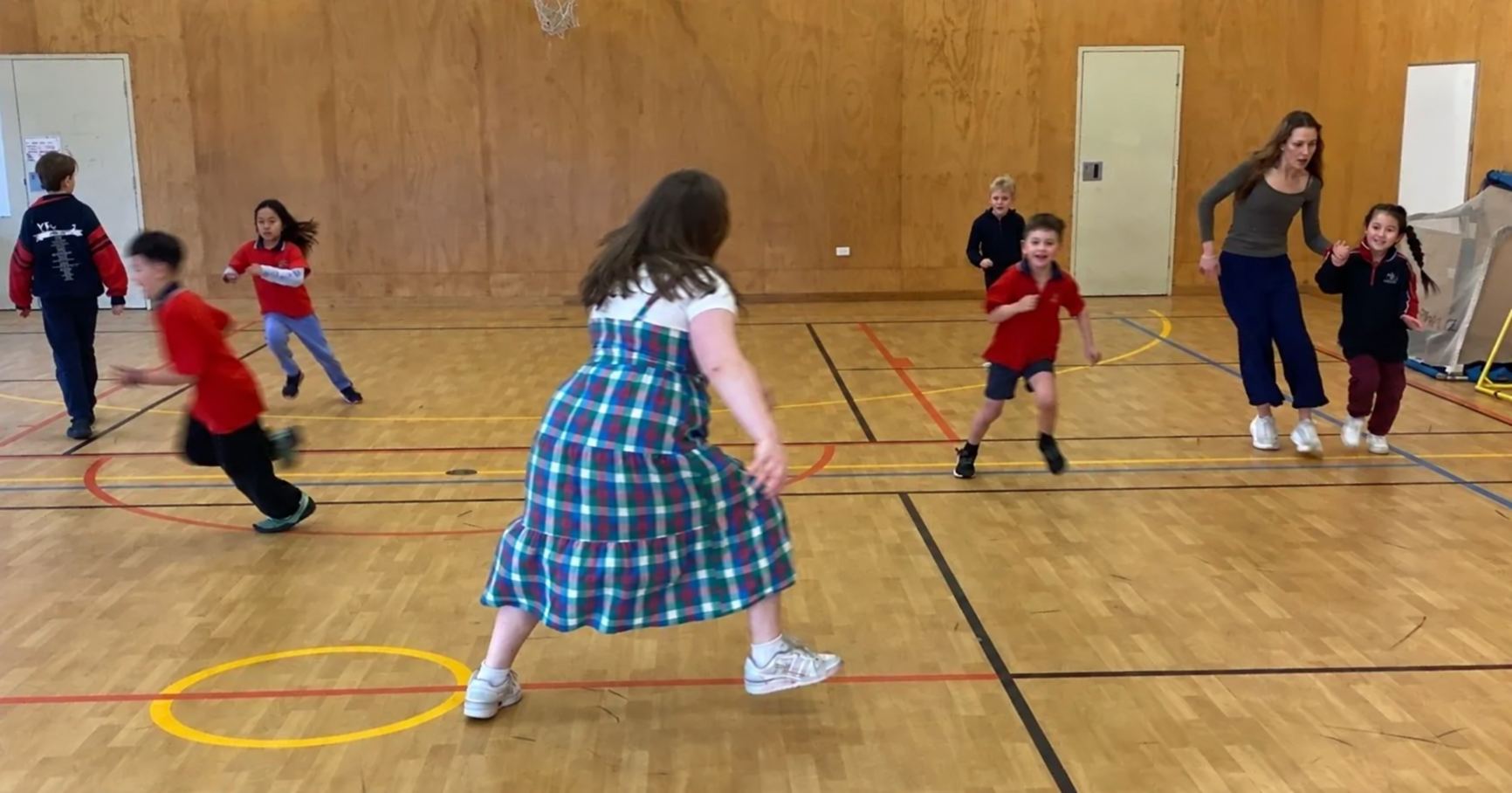Children playing and running in a gymnasium with a woman supervising.