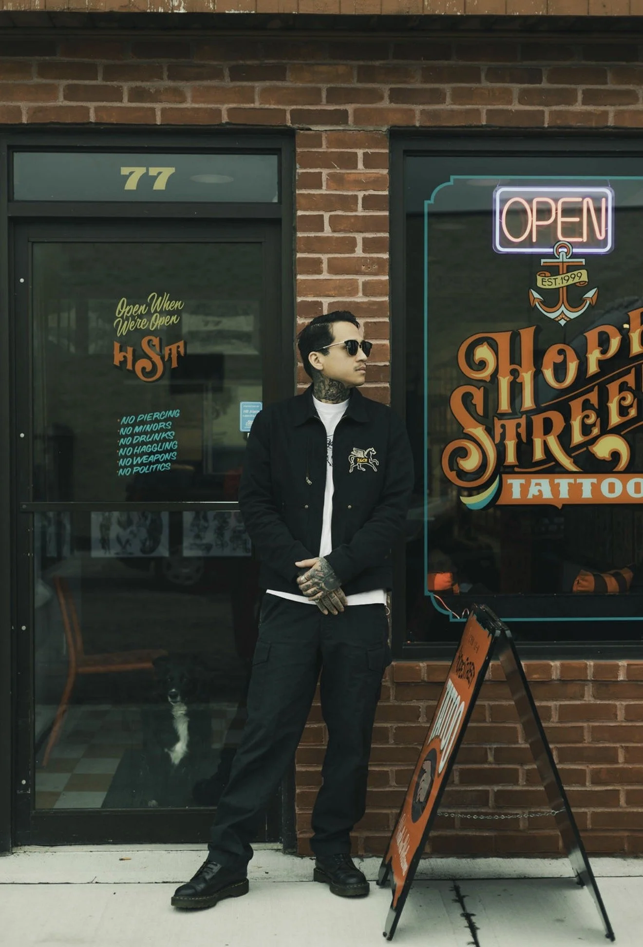 A man with tattoos, sunglasses, and dark clothing standing outside a tattoo shop with a brick wall background, a sign that says 'OPEN,' and a small chalkboard sign on the sidewalk.