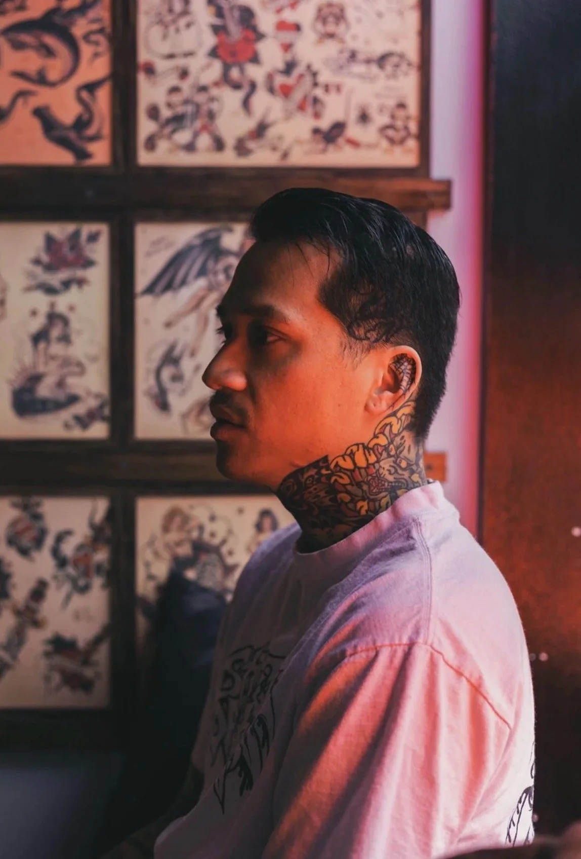 A young man with tattoos on his neck and ear, wearing a white T-shirt, looking to the left, with tattooed artwork on the wall behind him.