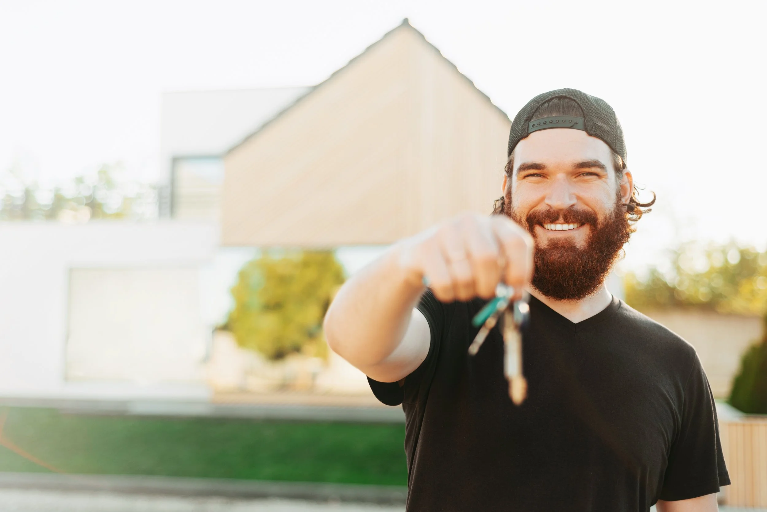 Man with a beard and baseball cap smiling and holding out keys outside in sunlight.
