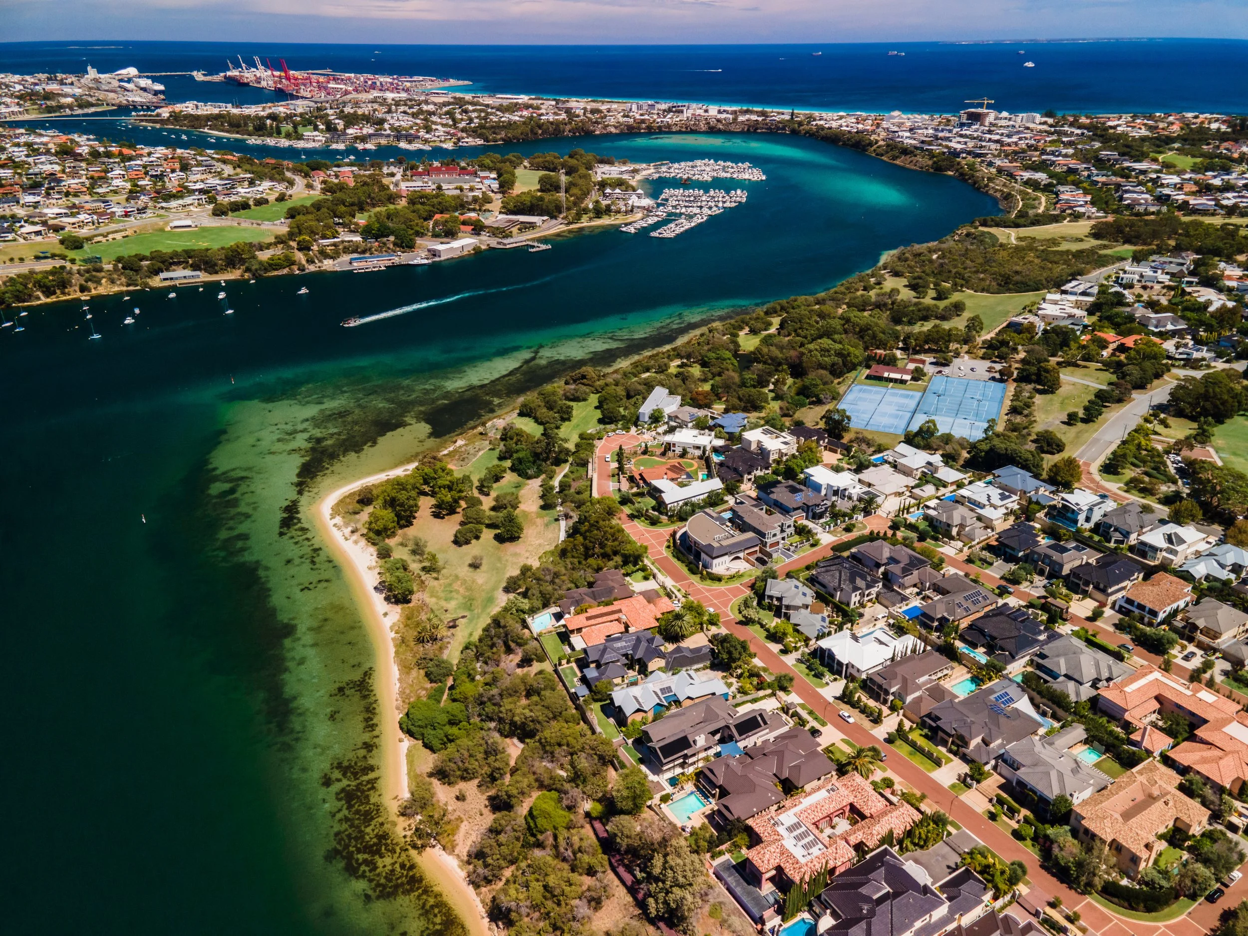 Aerial view of a coastal residential neighborhood with houses, tennis courts, and a beach along a greenish waterway with boats and a motorboat speeding. In the background, a harbor with cranes and ships, and the ocean.