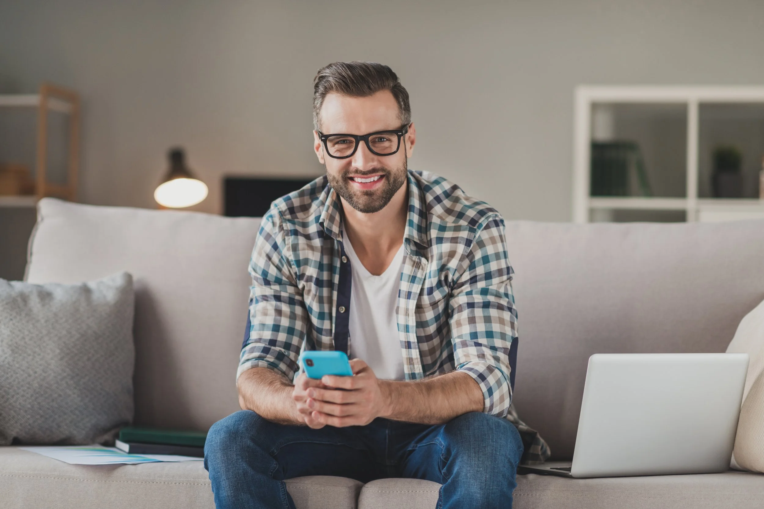 Smiling man with dark hair, beard, and glasses sitting on a beige couch using a smartphone, with a laptop and documents nearby in a cozy living room.
