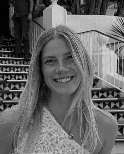 A woman with long blonde hair smiling at the camera in an outdoor setting with stairs and plants in the background.