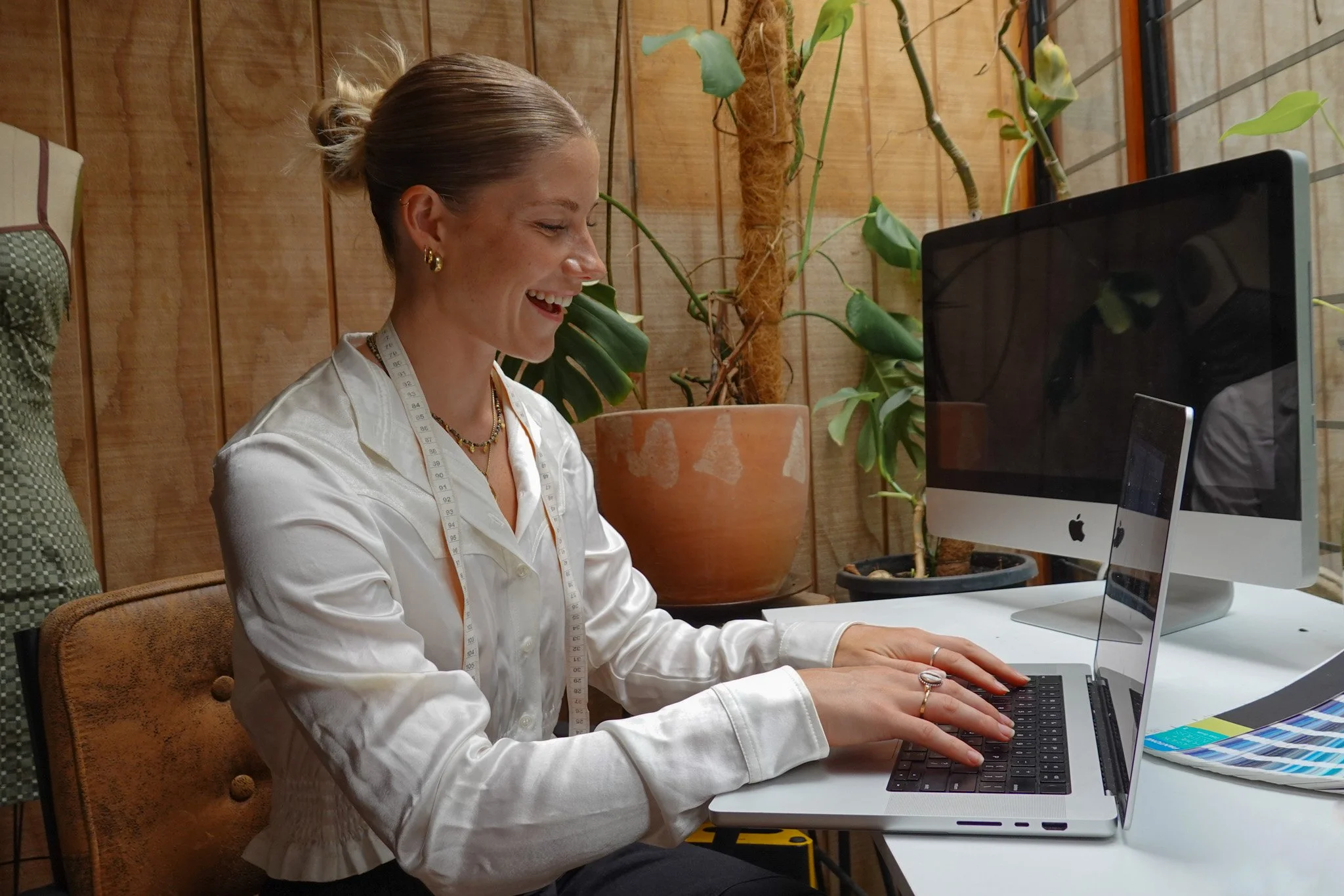 A fashion designer with blonde hair in a bun, wearing a white satin blouse and jewelry, sitting at a desk typing on a laptop in a cozy, plant-filled office space with wooden walls. Designing new prints and clothing for clients.