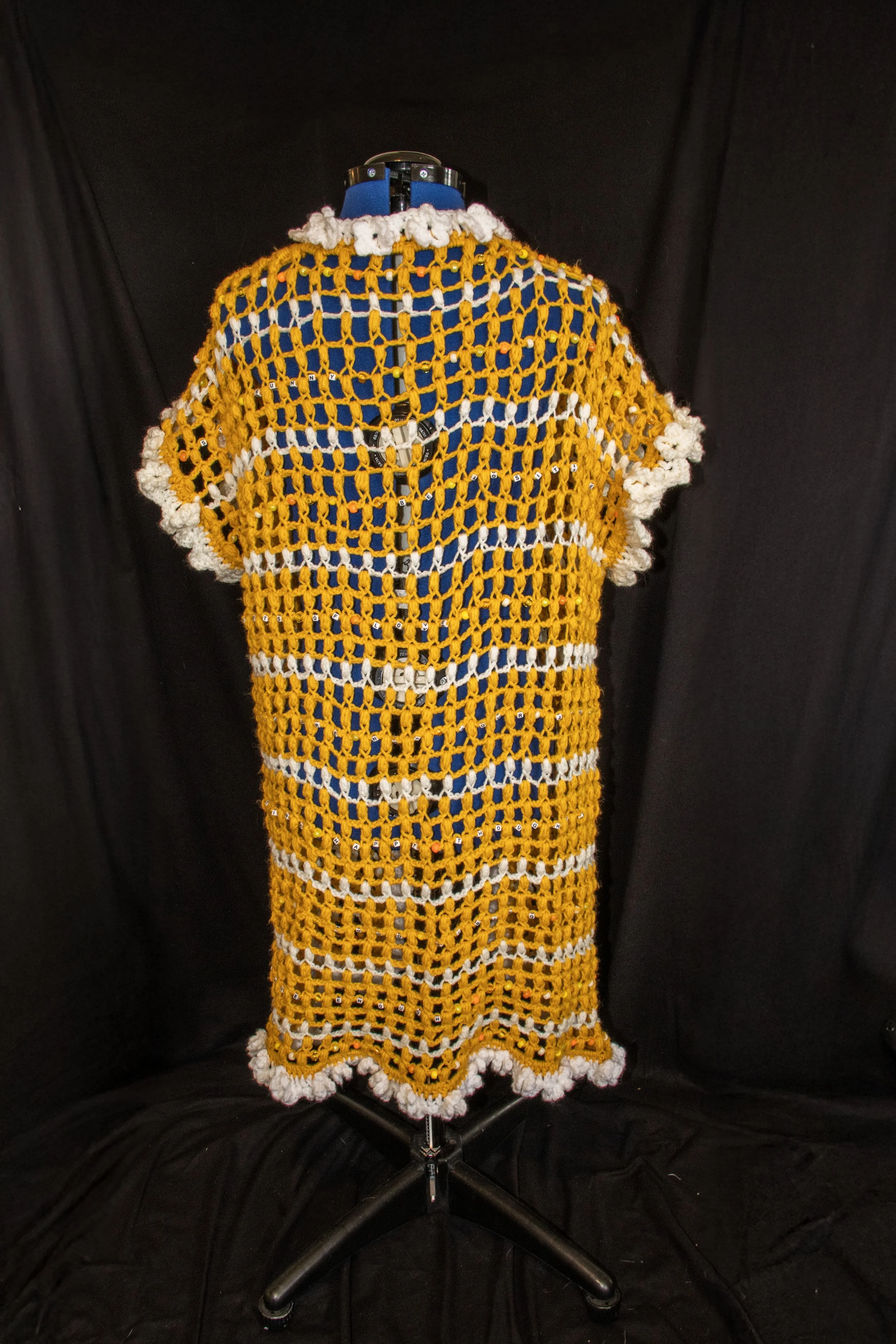 Back view of a mannequin dressed in a yellow, and white crochet cardigan with a floral edge, set against a black background.