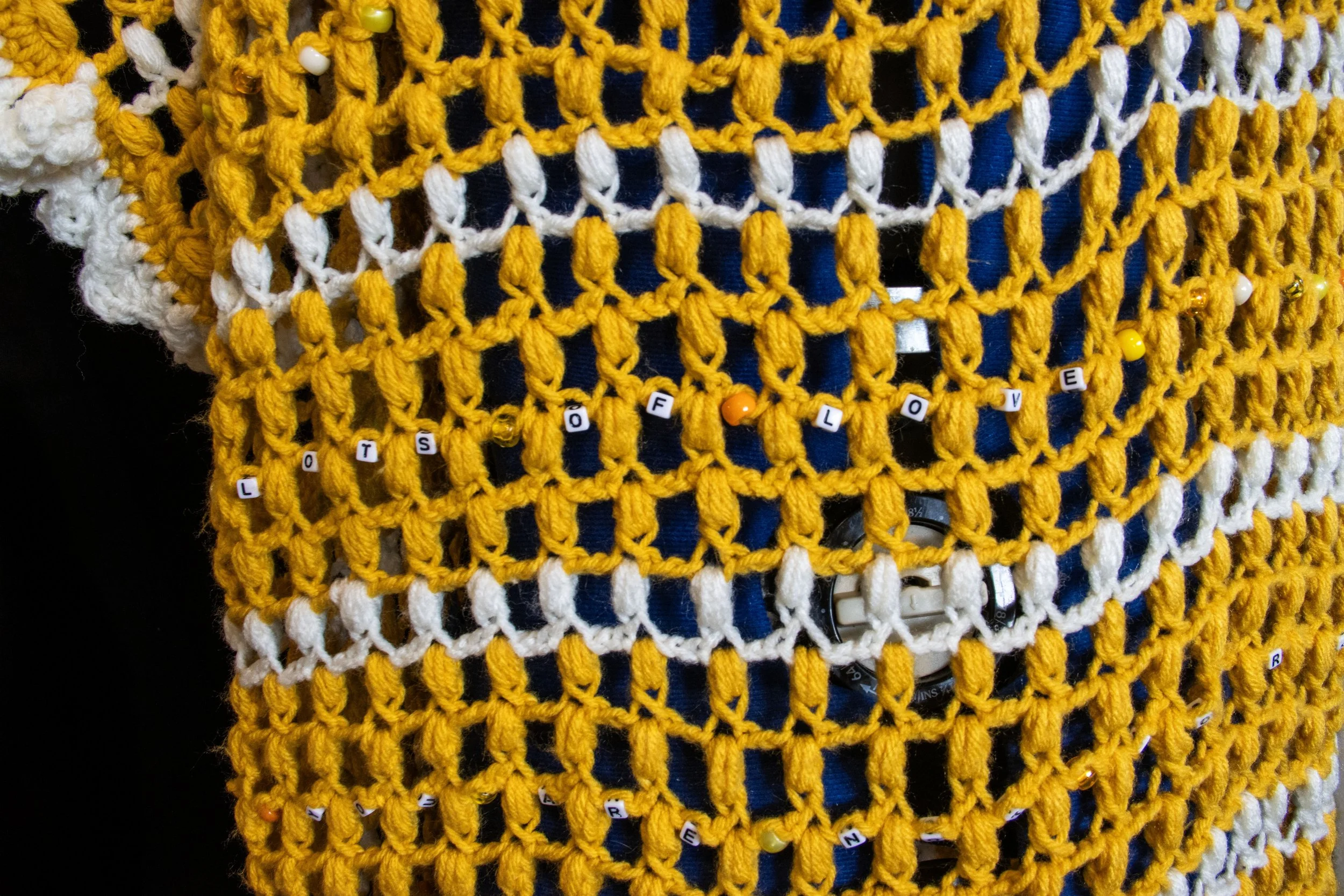 A yellow and white crocheted fabric with small letter beads attached, spelling out "LOTS OF LOVE." 