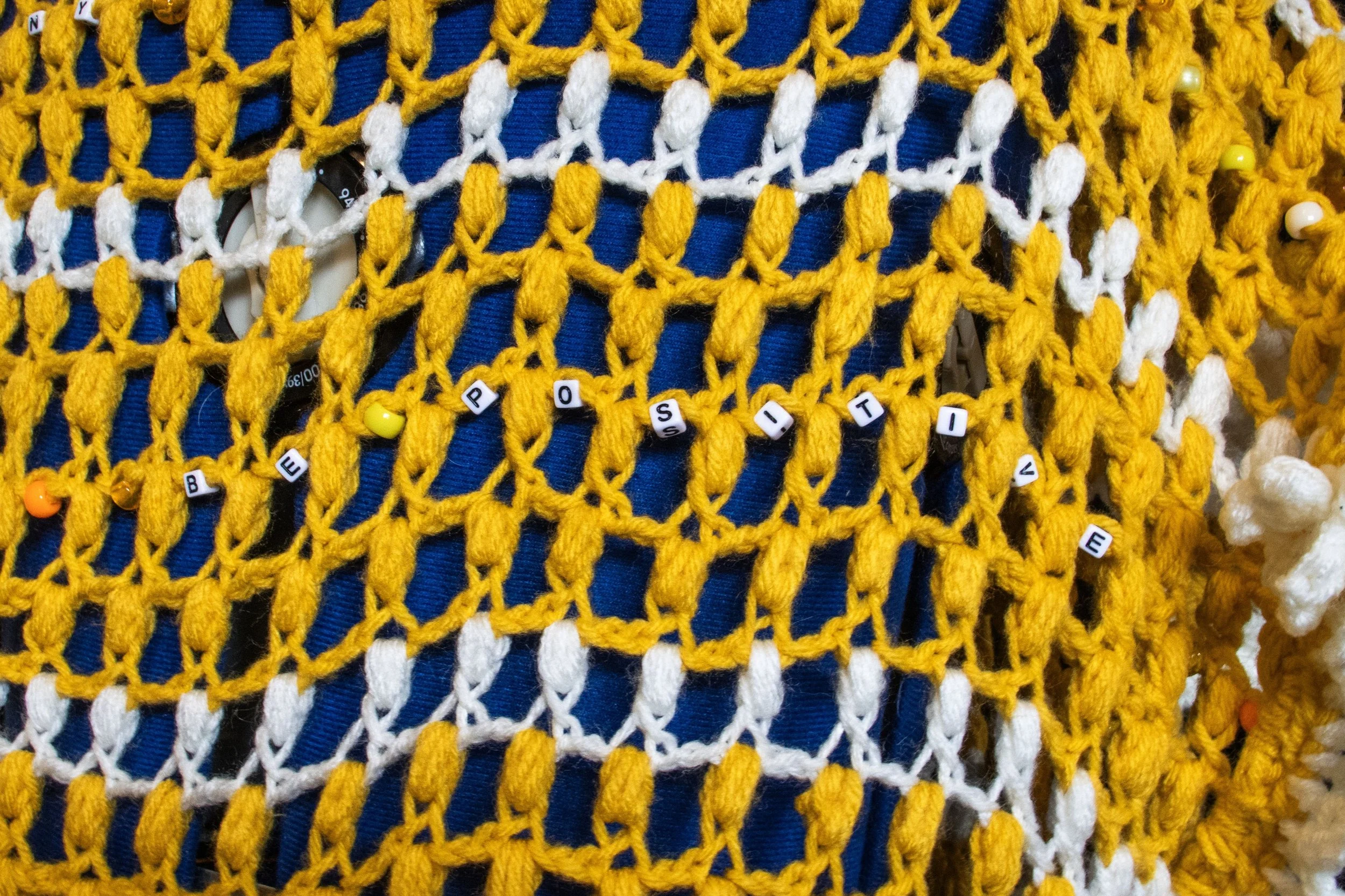 Close-up of a yellow and white crochet project with beads spelling out 'BE POSITIVE'