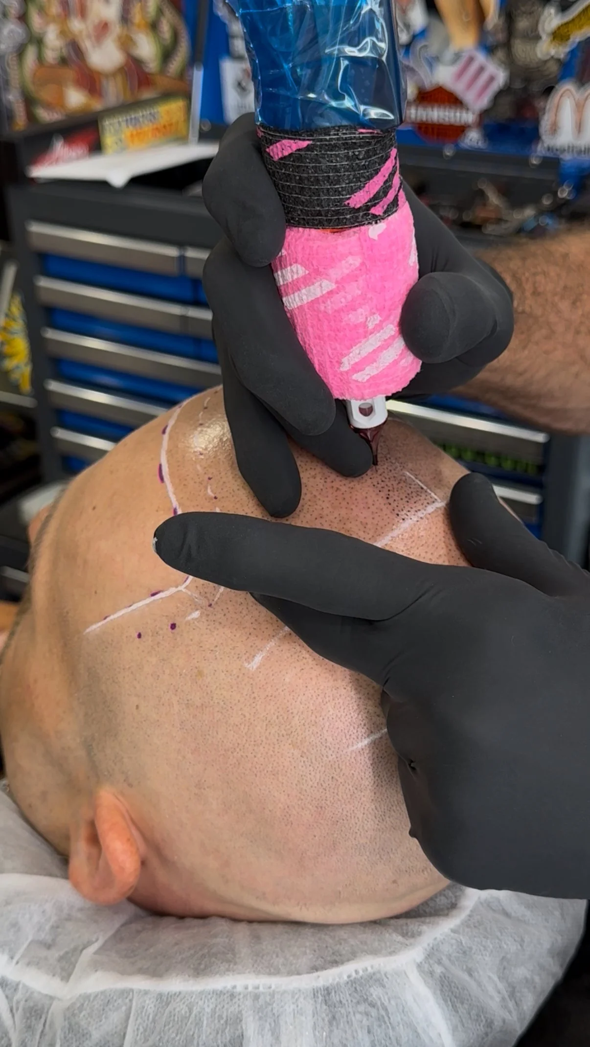 A person in black gloves is marking a man's shaved head with white chalk, preparing for a tattoo or micropigmentation procedure, in a workshop or tattoo studio.