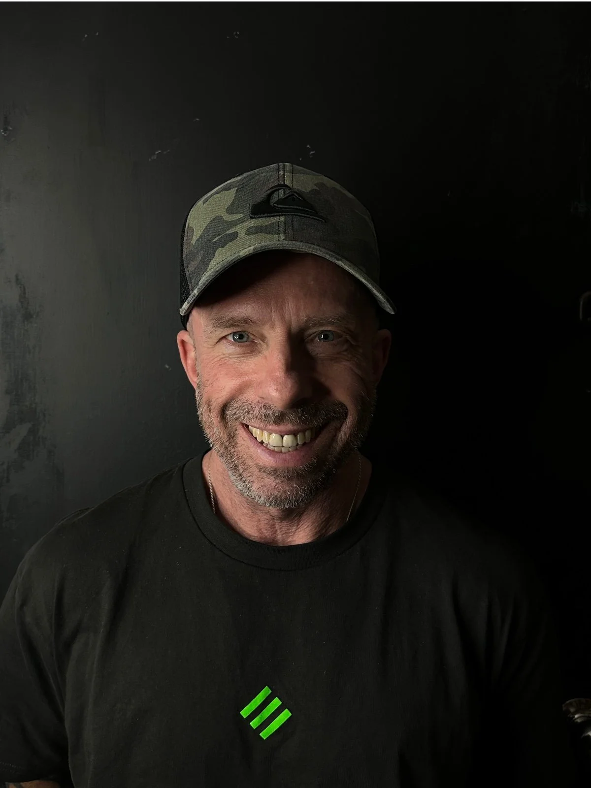 A smiling middle-aged man with a beard wearing a camo baseball cap and a black t-shirt, standing against a dark background.