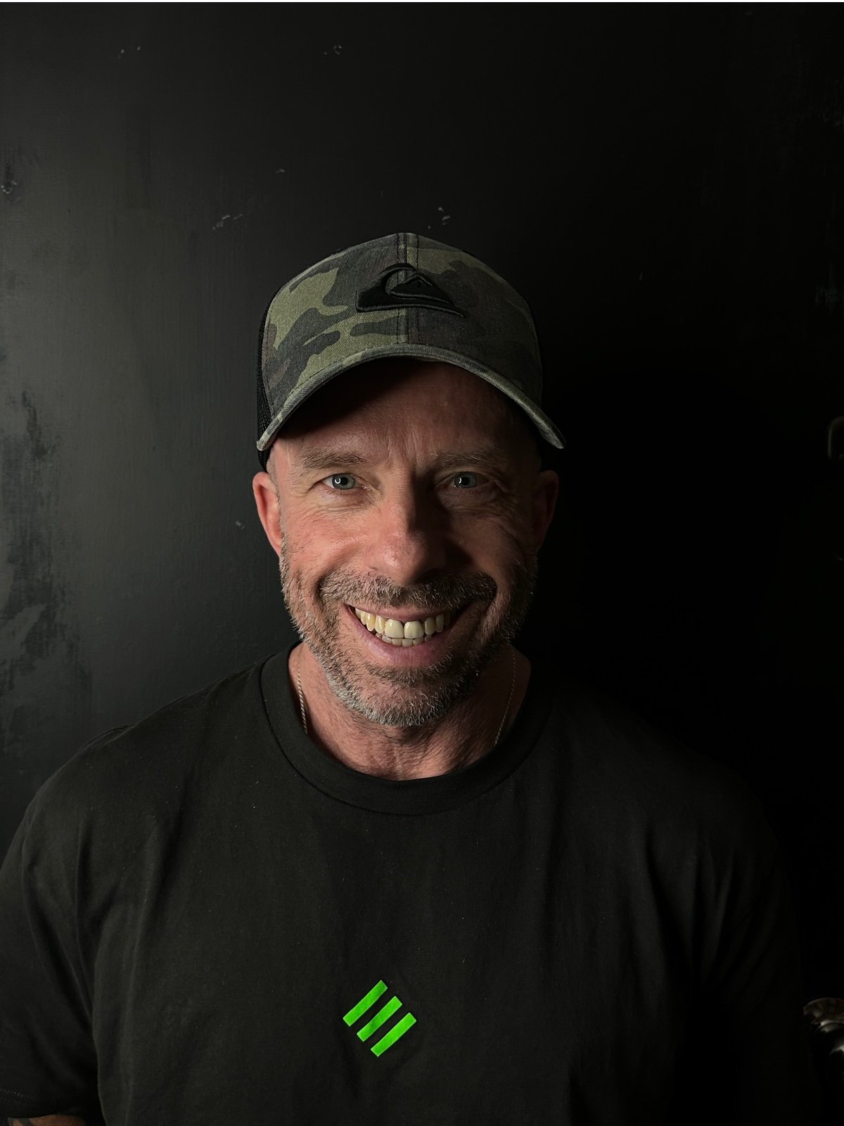 A smiling man with a beard and blue eyes wearing a camouflage baseball cap and a black t-shirt with a green logo on it, standing against a dark wall.