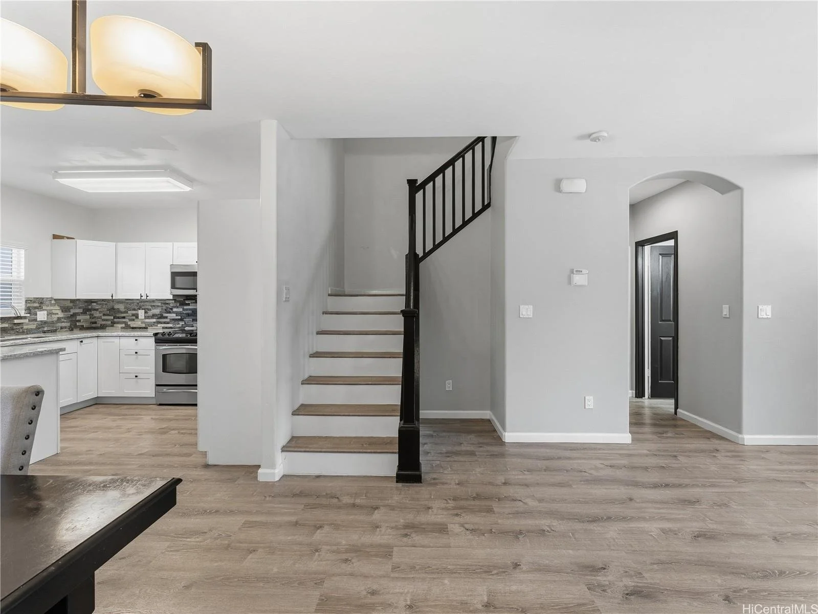 Interior of a modern house with a staircase, open kitchen with white cabinets, gray tile backsplash, stainless steel appliances, light wood flooring, and light gray walls.