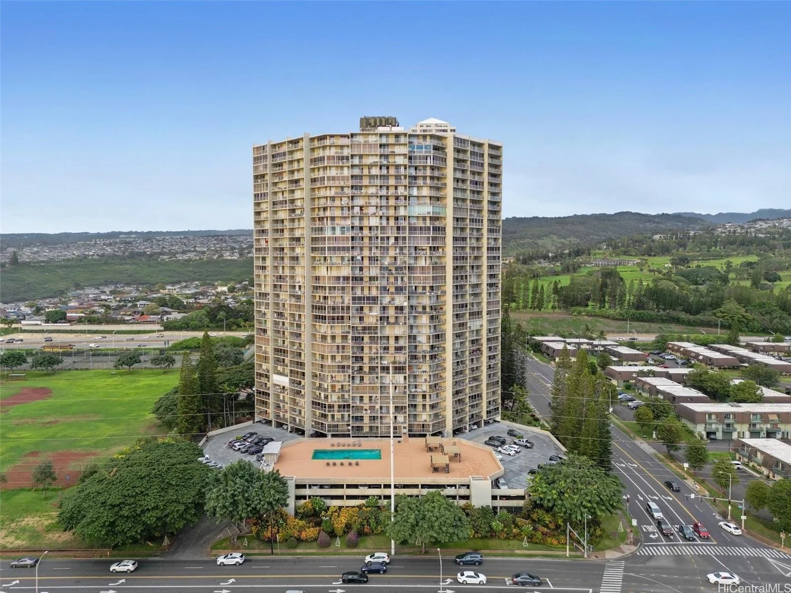 Tall residential building with many balconies, surrounded by greenery, with a parking lot and a swimming pool area on the lower level, and mountains in the background under a clear blue sky.