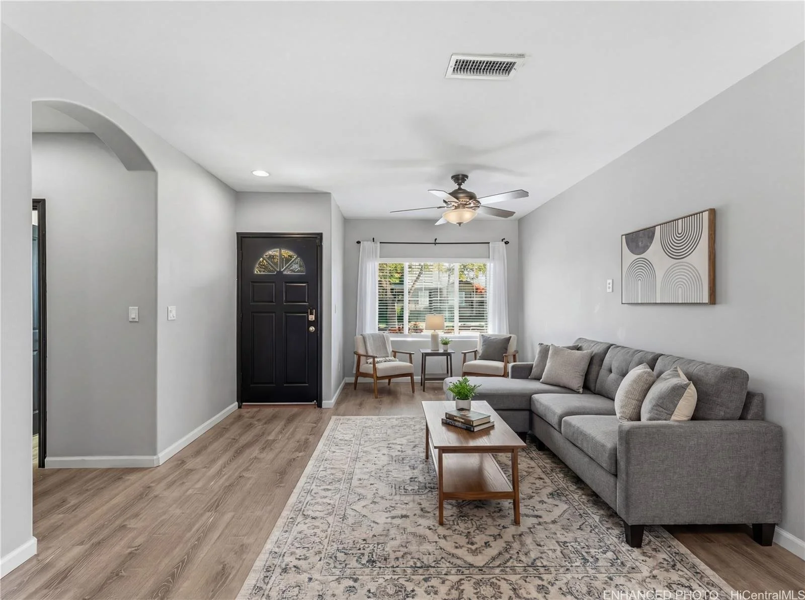 Living room with a gray sectional sofa, wooden coffee table, patterned rug, armchairs near a window, and a wall art piece.