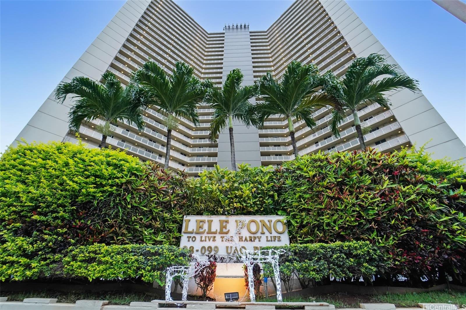Low-angle view of a tall modern apartment building with balconies, surrounded by lush green plants and palm trees, with a sign that reads "Lele Ono to Live Is to Happy Life" at the entrance.
