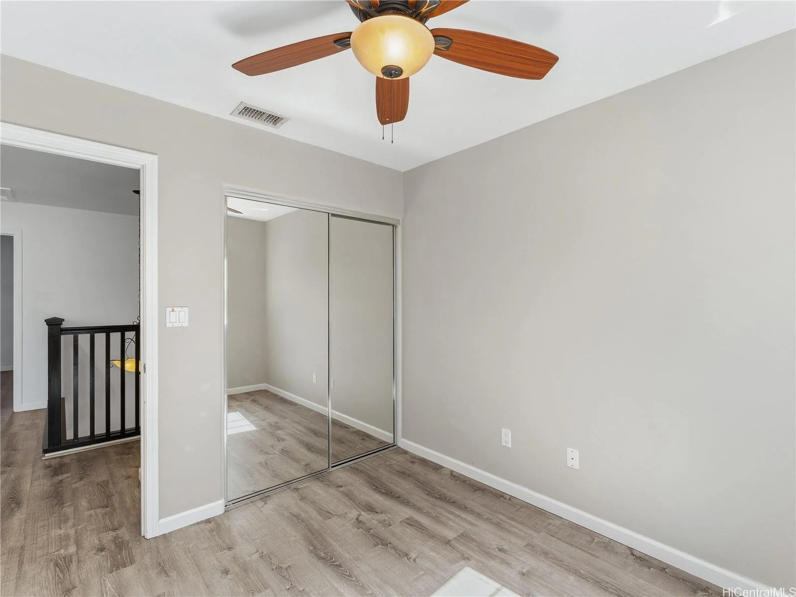 Empty beige room with hardwood floors, a ceiling fan with wooden blades, and a mirrored closet door.