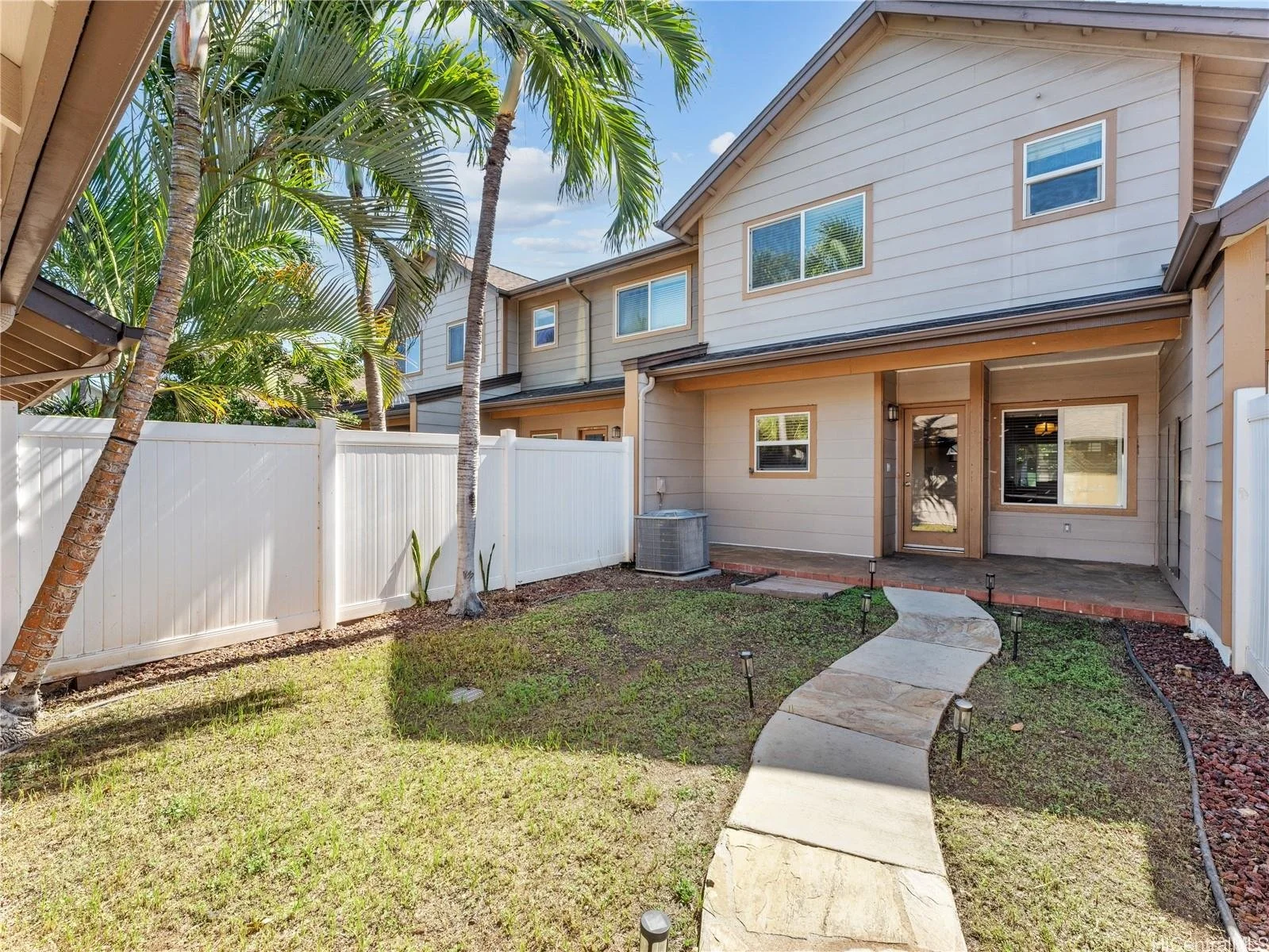 Backyard of a modern townhome with a curved stone pathway, small lawn, tall palm trees, surrounded by a white fence, under a blue sky with some clouds.