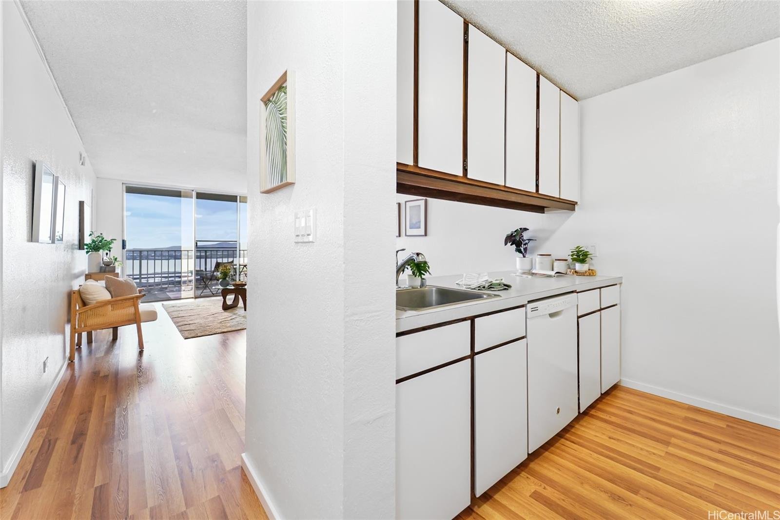 View of an open living room and kitchen in an apartment with wood floors, white walls, and a balcony with a view of the water.