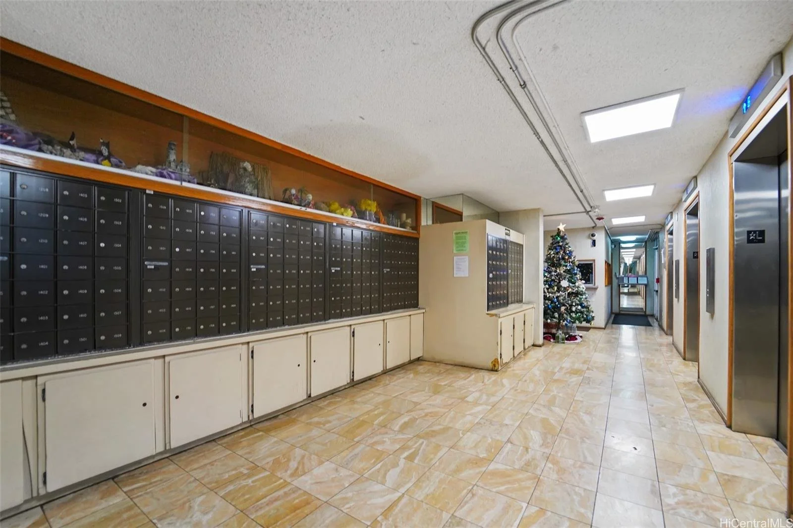 Inside a lobby with a row of black mailboxes on a wall, a Christmas tree decorated with ornaments, a door at the end, and elevators on the right