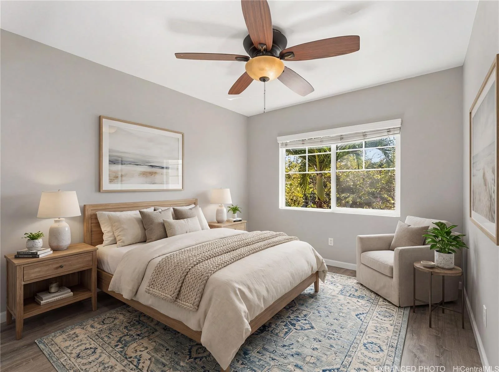 Bright bedroom with a wooden bed, surrounded by two nightstands, lamps, and decorative pillows. A window shows green trees outside, and there is a beige armchair with a small table and plant next to it. The room has light-colored walls, a ceiling fan, and a patterned rug.
