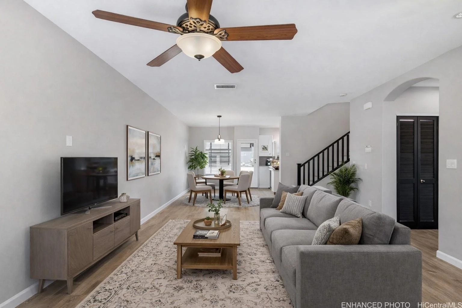 Open-concept living and dining area with gray sofa, wooden coffee table, TV on console, artwork on wall, dining table with four chairs, potted plant near window, and staircase with black railing