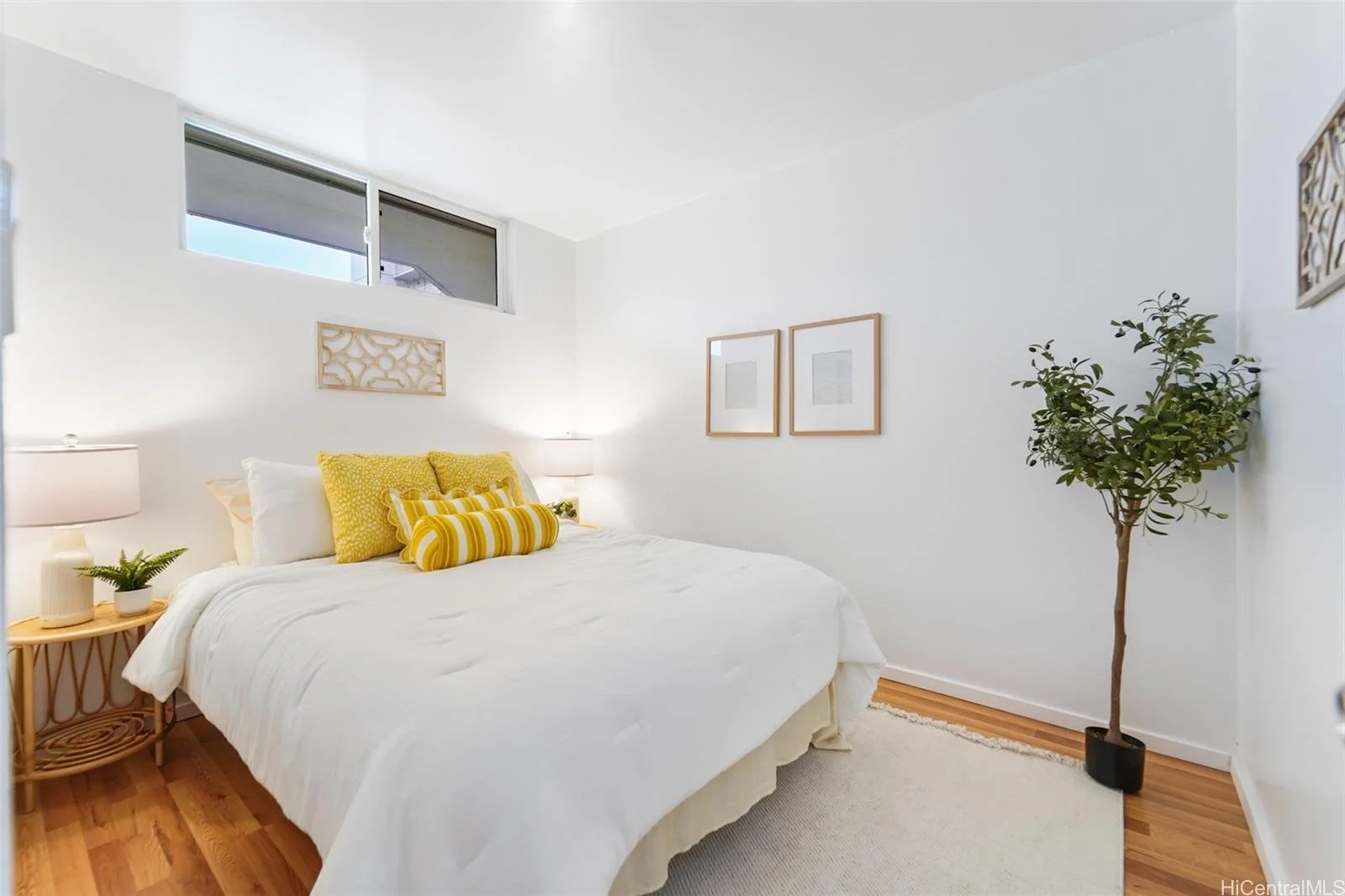 A bedroom with white walls and bedding, yellow decorative pillows, a wooden side table with a small plant, a white lamp, two framed pictures on the wall, and a tall potted plant in the corner.